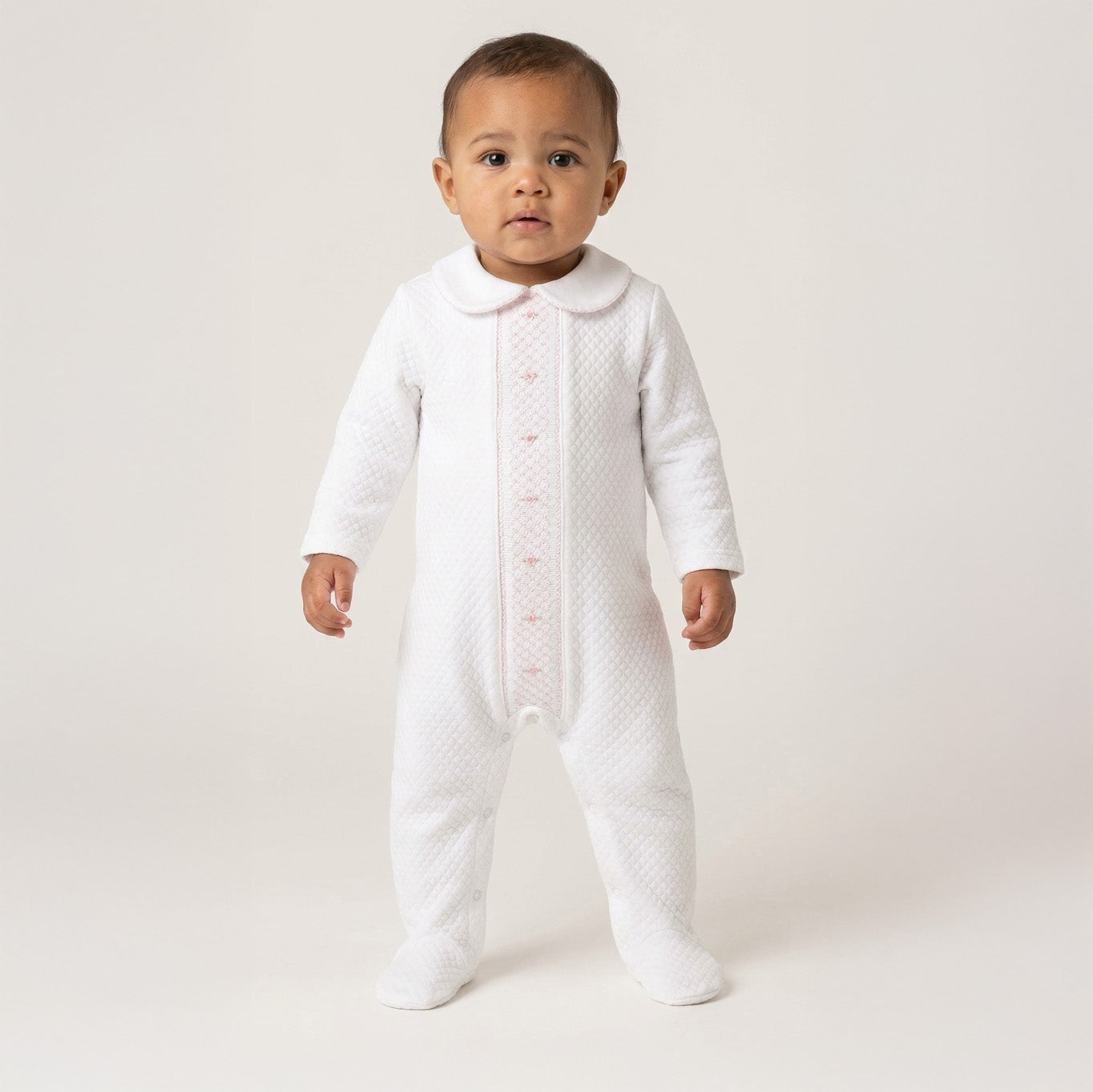 Baby standing in the white quilted Chéris Mademoiselle babygrow with pink floral embroidery and smocked detail.